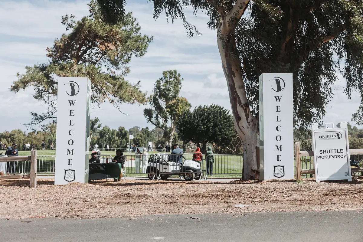 Twin WELCOME pylons at the spectator entrance to Palos Verdes Golf Club, shuttle carts waiting on the gravel behind them