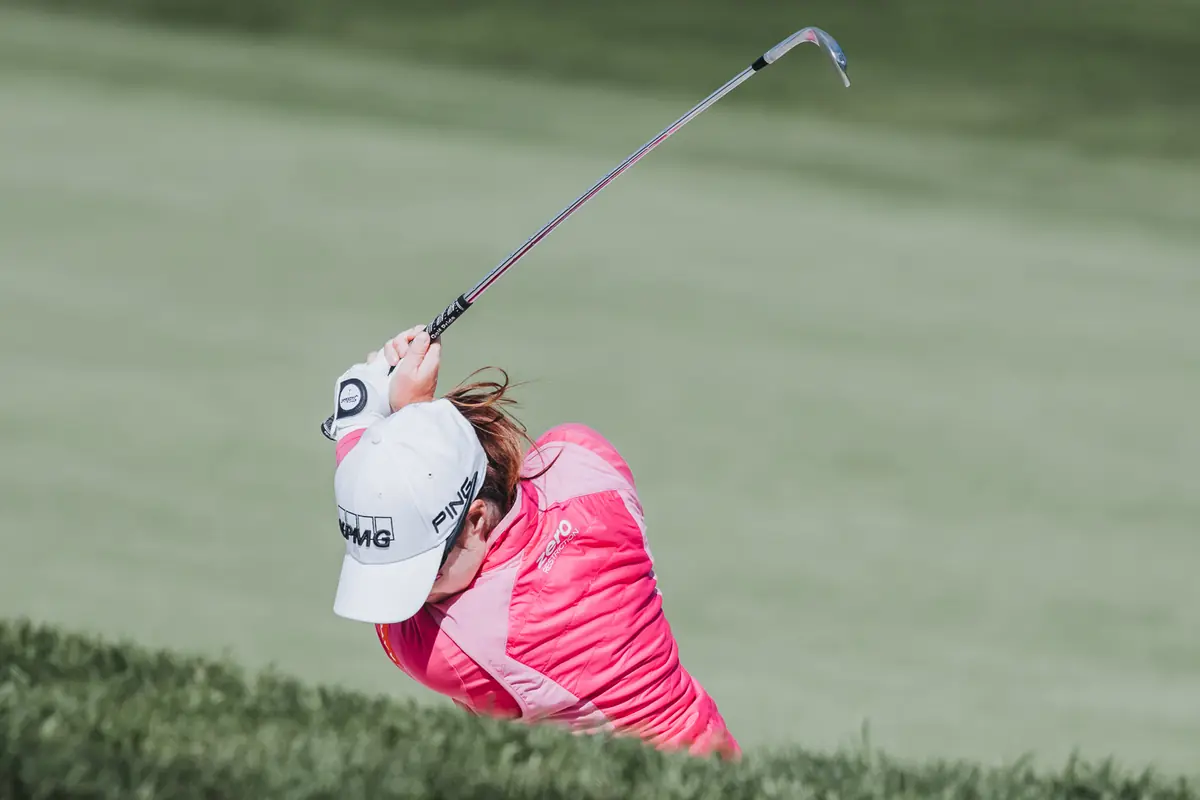A player in pink following through over a clipped hedge line, club wrapped behind her shoulders