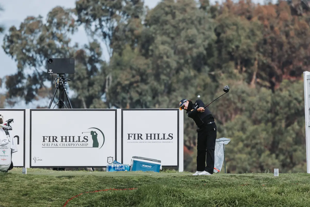 A tee shot framed between the Fir Hills sponsor boards and a television camera on the tee