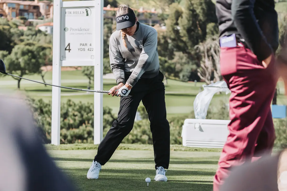 A tee shot seen between two spectators standing at the ropes, the peninsula falling away behind