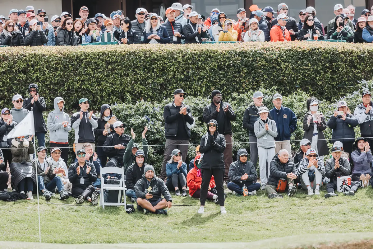Spectators stacked several rows deep behind a clipped hedge, phones up, watching the green