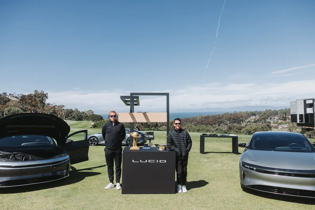 Two Lucid sedans displayed on the grass above the ocean with the trophy on a plinth between them