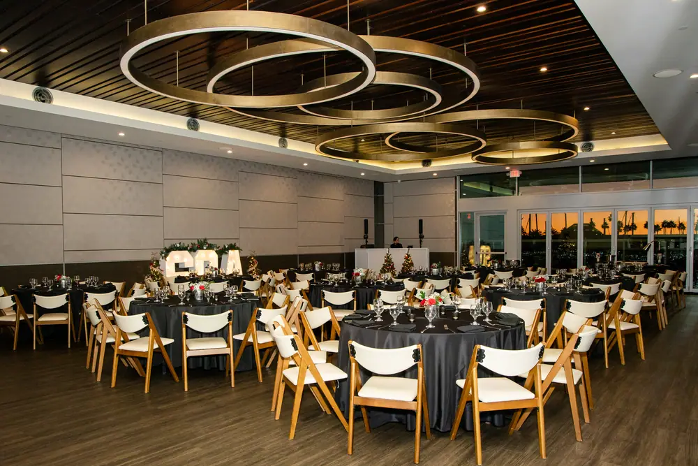 The Sky Level room at Shade Hotel Redondo Beach set for SQA Services' 30th anniversary dinner — black-linen rounds, pale wood chairs and gold ring chandeliers with the sunset in the windows