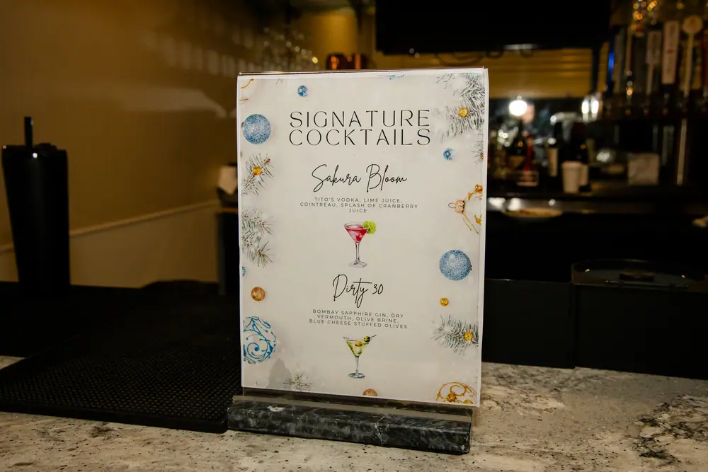 Signature cocktail card on the bar listing the Sakura Bloom and the Dirty 30 at the SQA Services holiday party
