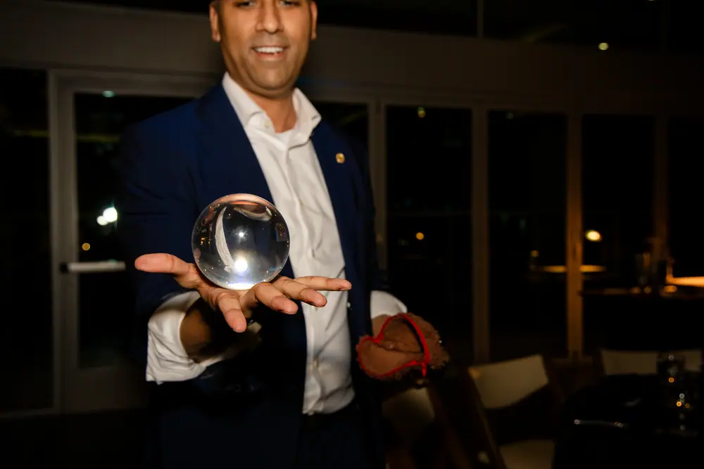 A close-up magician balancing a clear crystal ball on an open palm, Redondo Beach harbour lights behind him